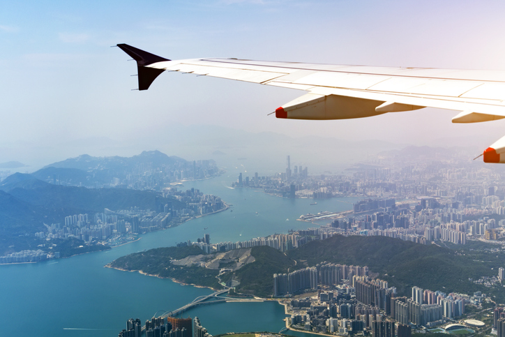 From a window seat, the airplane wing is visible as it flies above Hong Kong. The coastline and city are below. The scene captures the vastness of the city and the hilly terrain