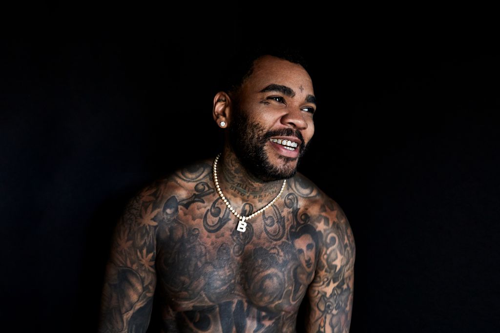 Tattooed man with beard smiling against a dark background, wearing a gold necklace with a pendant.