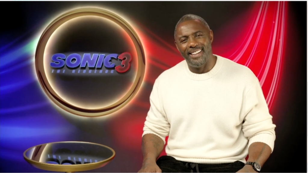 A smiling man in a white sweater standing in front of a "Sonic the Hedgehog 3" logo.