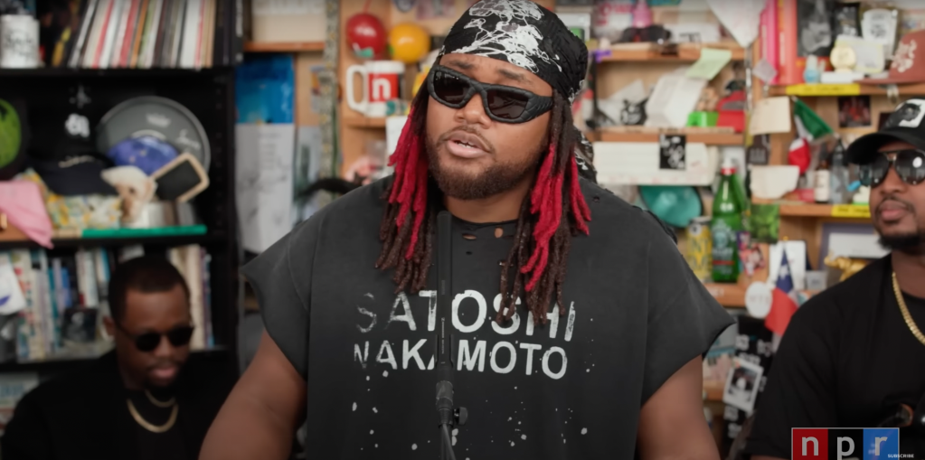 A person wearing sunglasses, a bandana, and a t-shirt that says "Satori Naki Moto" standing in a room filled with various objects and memorabilia.
