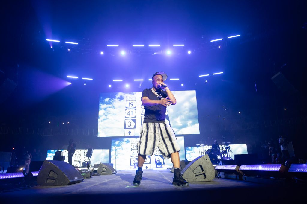 A performer on stage in a concert setting, wearing a hat and baggy pants, holding a microphone and surrounded by bright blue lighting and video screens.