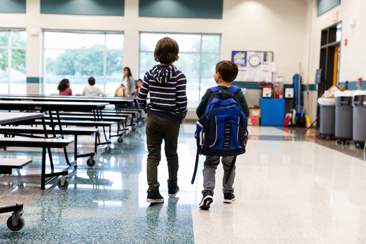 Two young boys walking down a school hallway with backpacks.