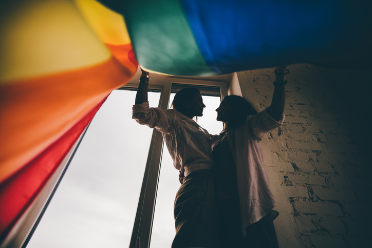 A silhouetted lesbian couple stands by a window, holding a vibrant rainbow flag above them.