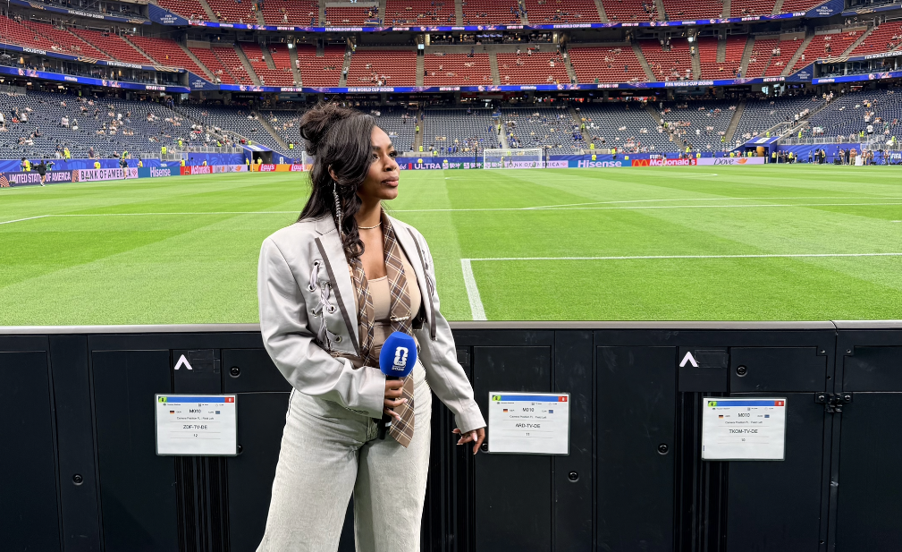 A woman stands on a soccer field in front of a large "HOUSTON" display at a sports stadium.