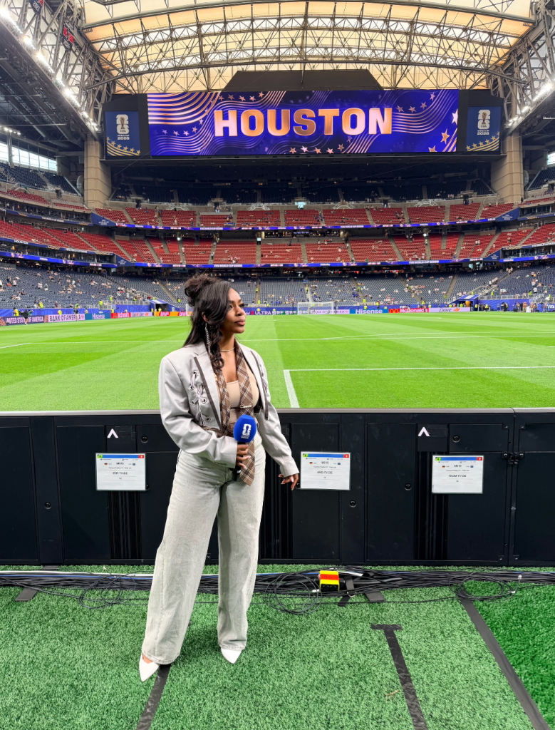 A woman stands on a soccer field in front of a large "HOUSTON" display at a sports stadium.