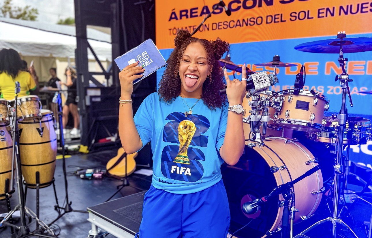 A smiling woman in a blue FIFA World Cup t-shirt holding a FIFA World Cup credential and standing in front of a stage with musical instruments.