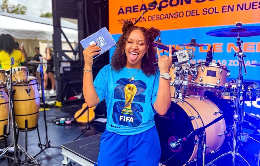 A smiling woman in a blue FIFA World Cup t-shirt holding a FIFA World Cup credential and standing in front of a stage with musical instruments.