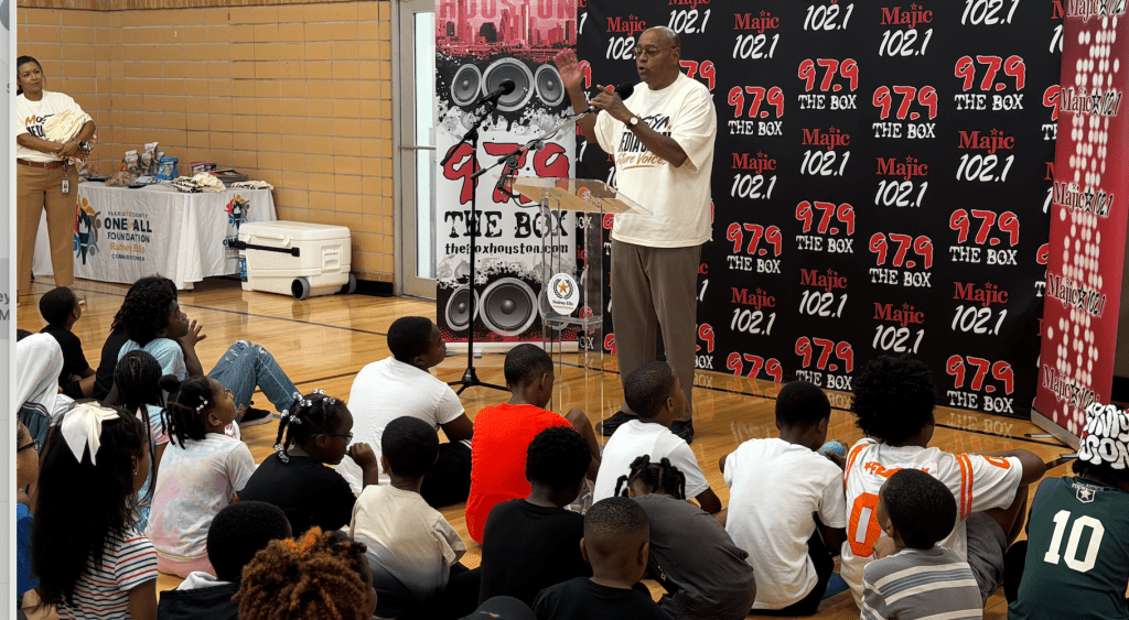 A group of people listening to a speaker in front of a backdrop with "The Box" and radio station logos.