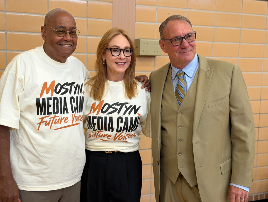 Three people smiling and wearing "Mostyn Media Camp" t-shirts in a hallway.