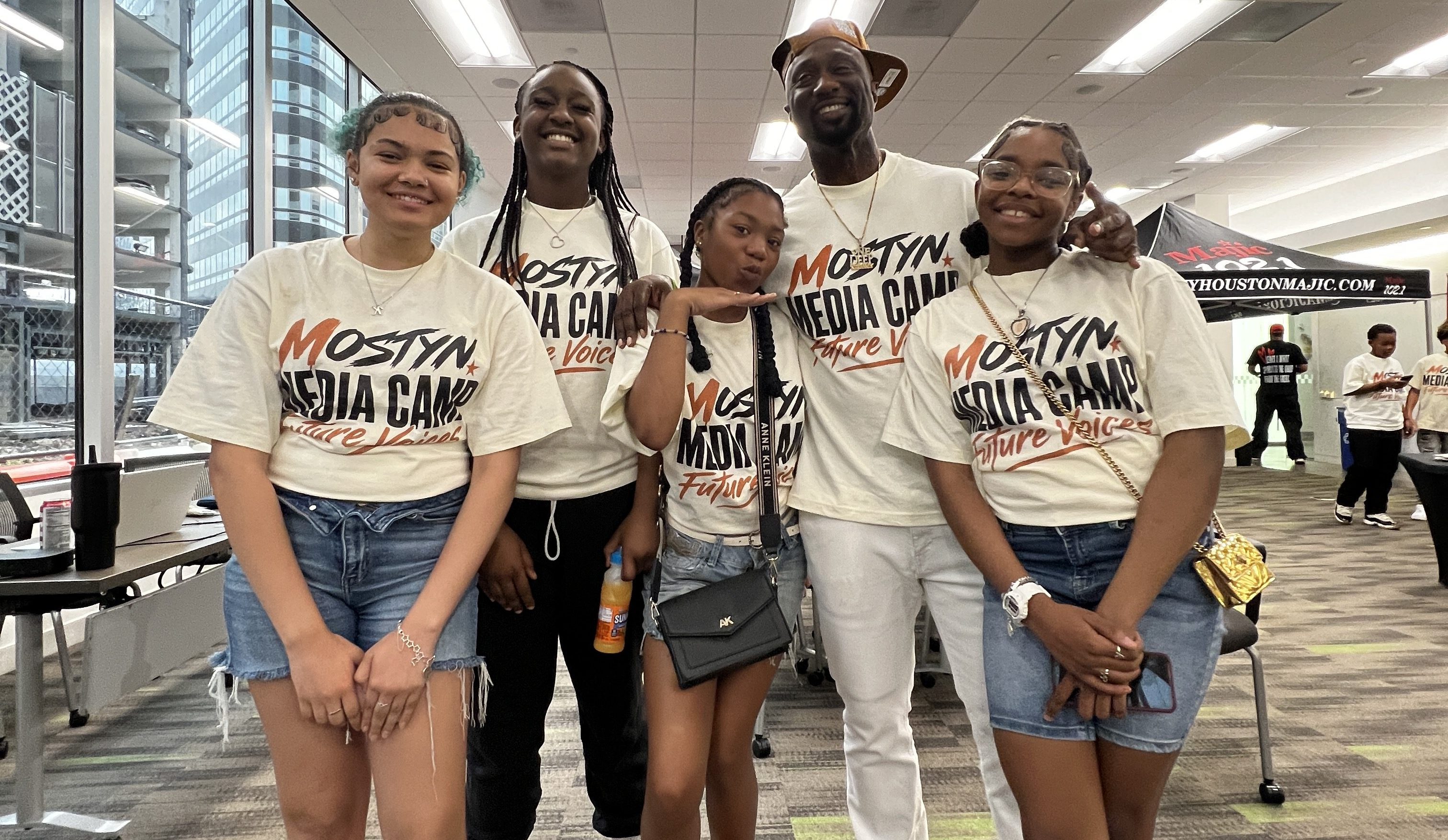 A group of five young people wearing t-shirts that say "Mostyn Media Camp" posing together in an indoor setting.