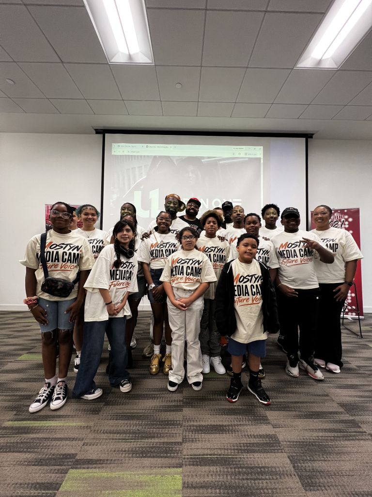 A group of people wearing t-shirts that say "Mosty Media Camp" posing together in a room with a projection screen.