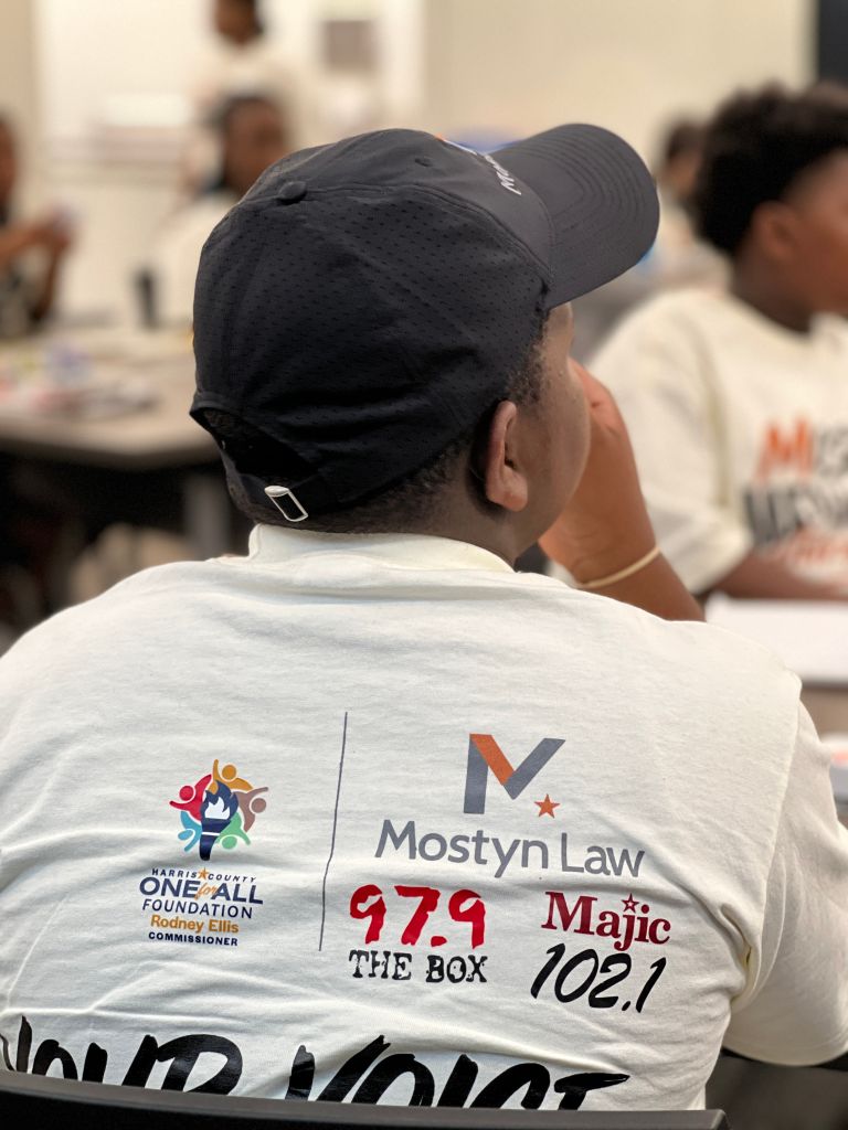 A person wearing a black hat and a white t-shirt with the text "Mostyn Law 97.9 The Box Magic 102.1" and the Harris County One Call Foundation logo.