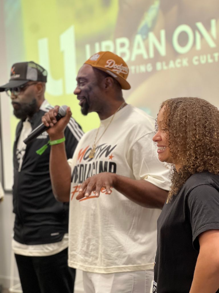 Three people on stage, one holding a microphone and wearing a "Dodgers" cap, another wearing a "Urban on" t-shirt, and a woman with curly hair smiling.