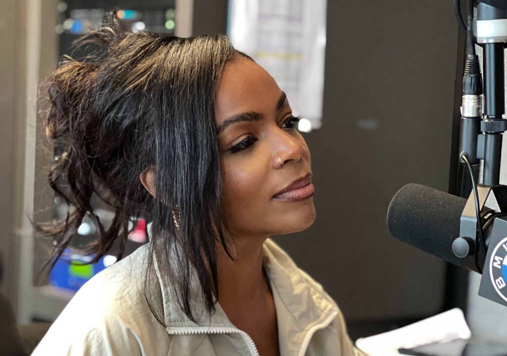 A woman with curly dark hair wearing a beige jacket sits at a desk in front of a microphone, appearing to be in a radio or podcast studio.