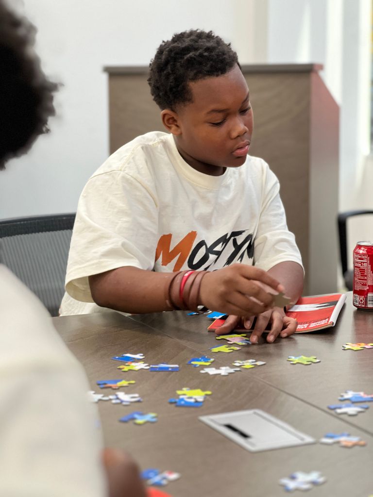 A young Black boy wearing a white t-shirt with "Monya" printed on it, sitting at a table and working on a puzzle.