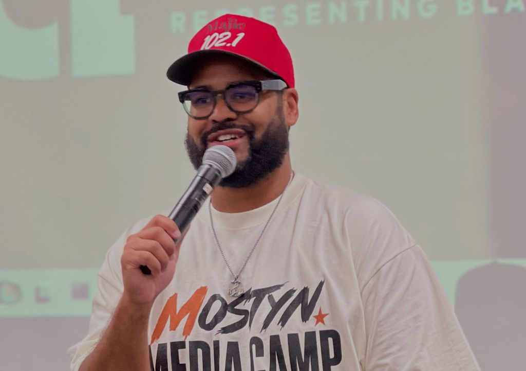 A man wearing a red cap and glasses holding a microphone and speaking at a podium with the "Urban Representing Black" logo visible.