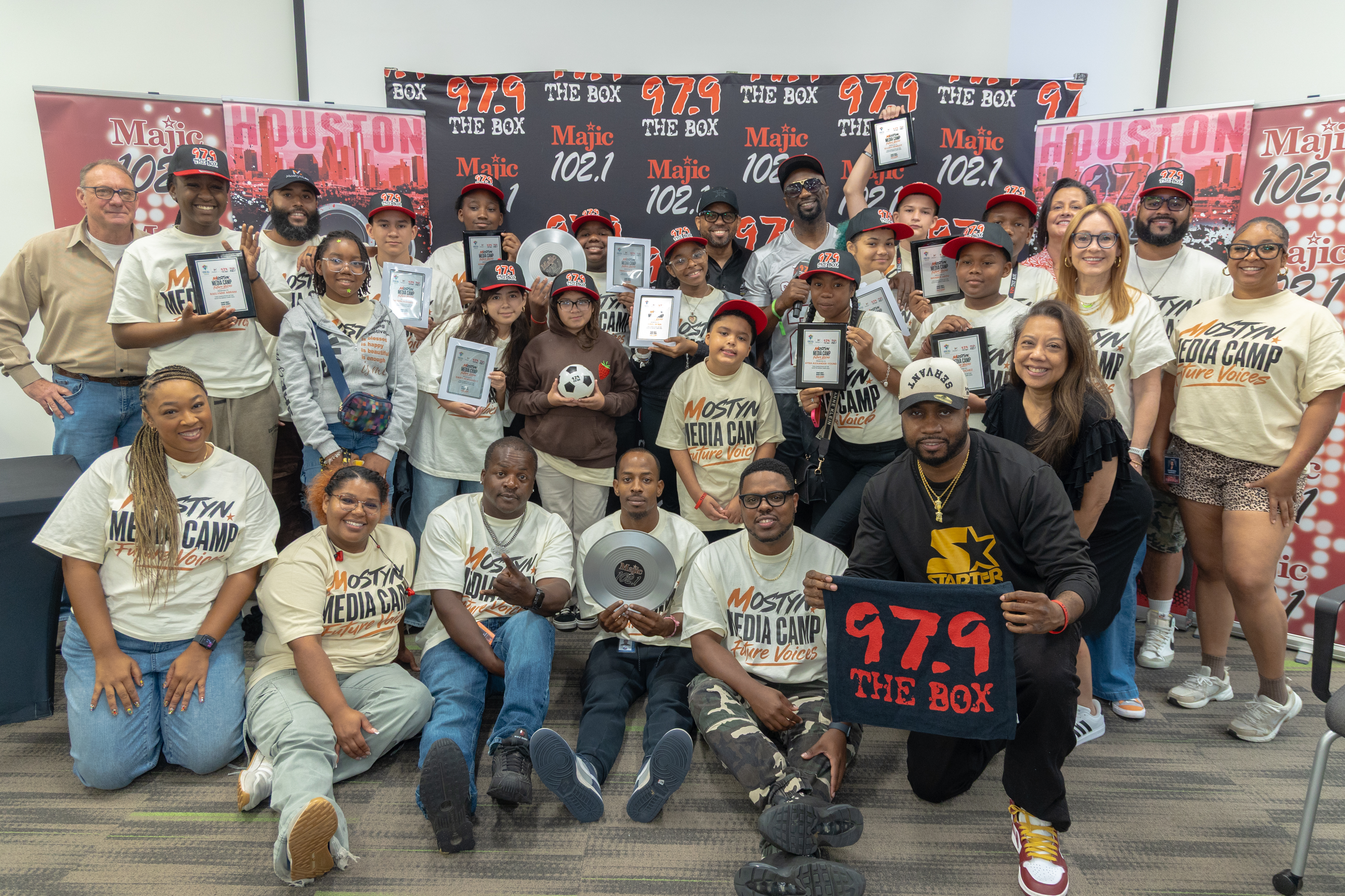 A group photo of people wearing "Majic 102.1" and "The Box 979" branded shirts, holding various media devices and awards, gathered in a radio station setting.