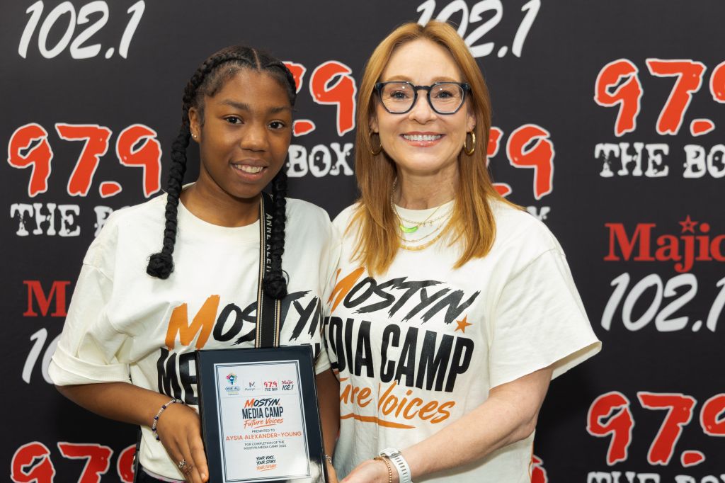 Two young women, one with braided hair and the other wearing glasses, standing in front of a radio station backdrop with the text "Moslyn Media Camp" and "The Voices".