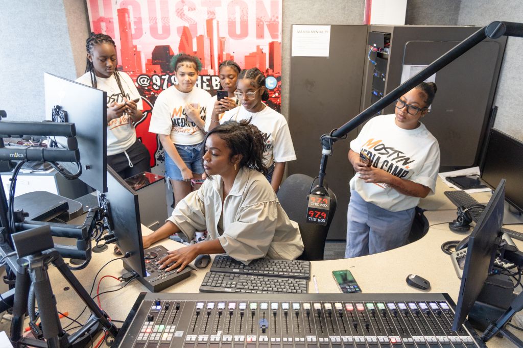 A group of young people working together in a radio studio, with audio equipment, computers, and a large "FUSION" sign on the wall.