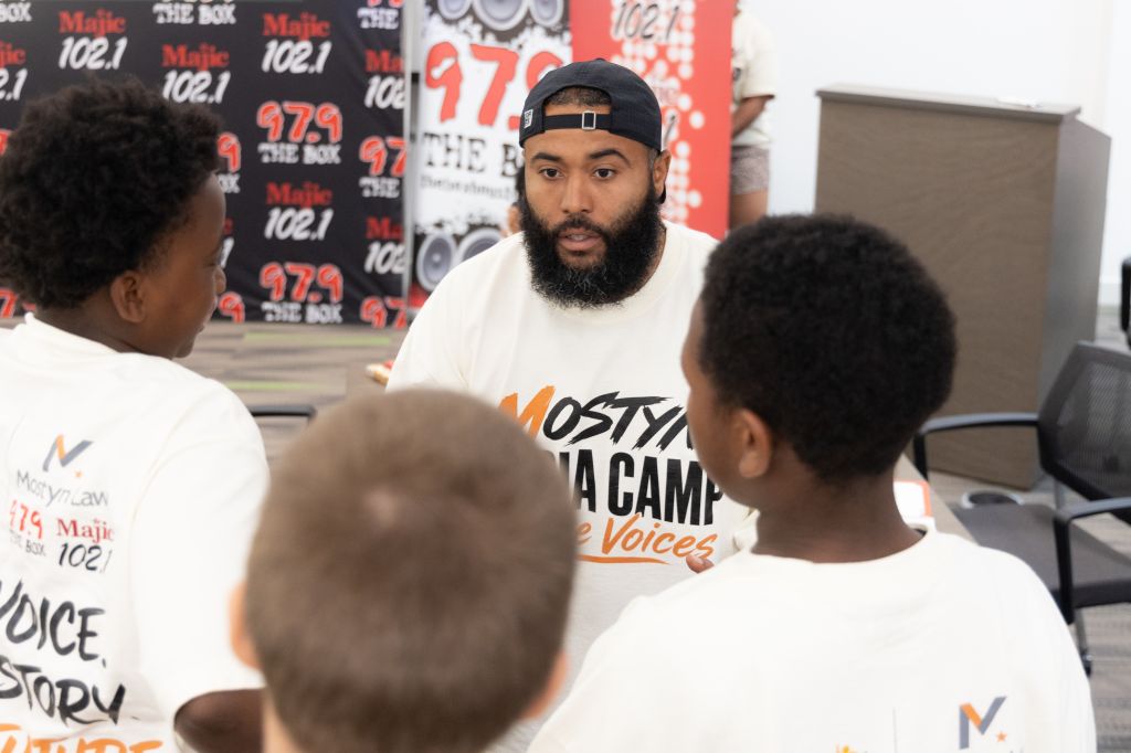 A man with a beard wearing a "Mostly Ya Camp" t-shirt speaking to two young boys in front of radio station branding.