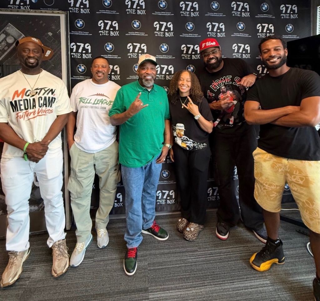 A group of five people posing together in front of a backdrop with "979 THE BOX" branding. The people are wearing casual clothing and appear to be at an event or promotional setting.