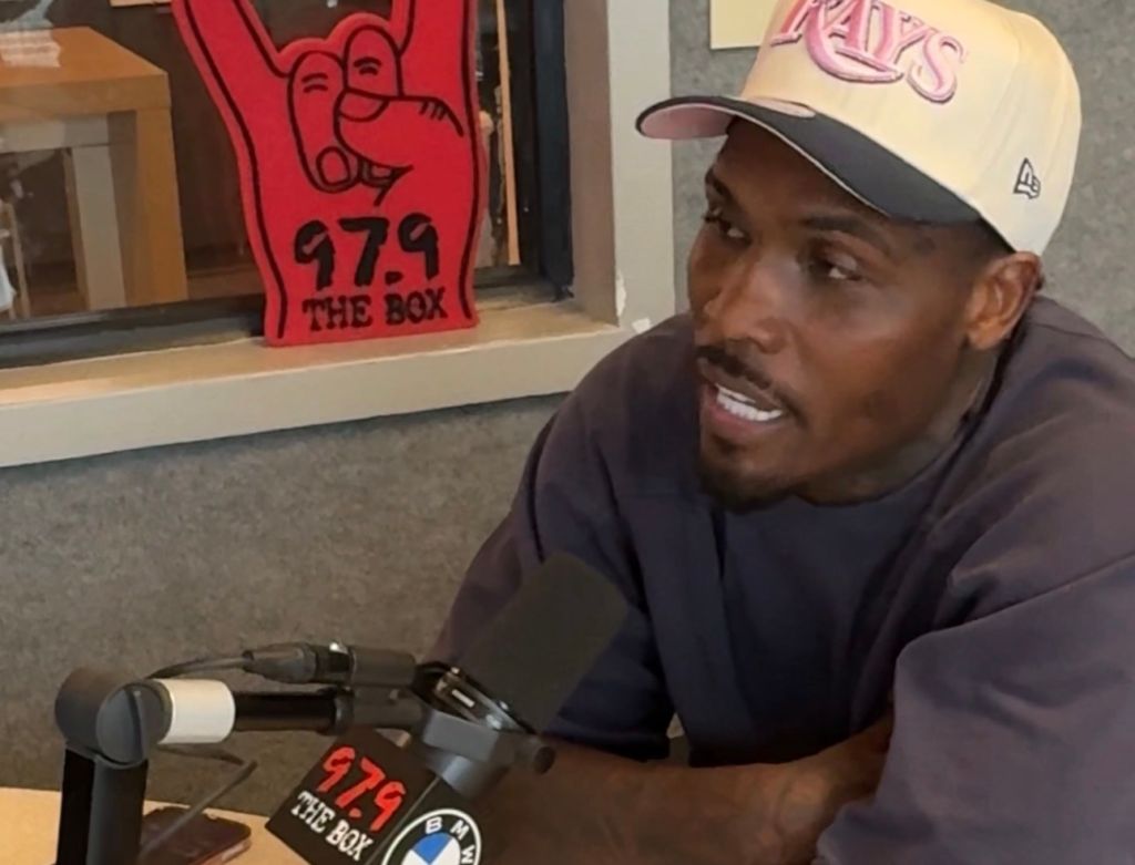 A man wearing a New Era cap and a black shirt is sitting in front of a radio station sign that says "979 THE BOX".