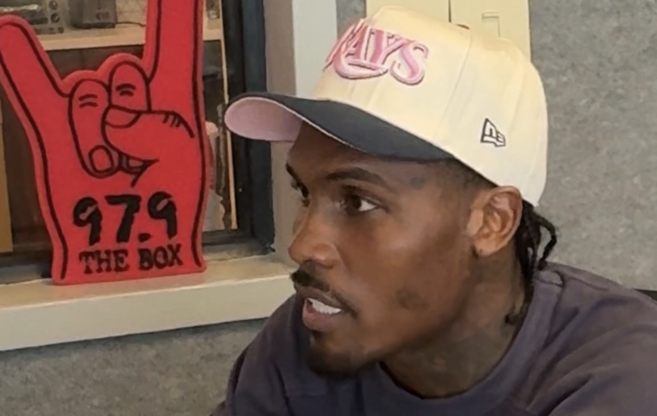 A man wearing a New Era baseball cap and a dark jacket, standing in front of a sign that says "97.9 THE BOX".