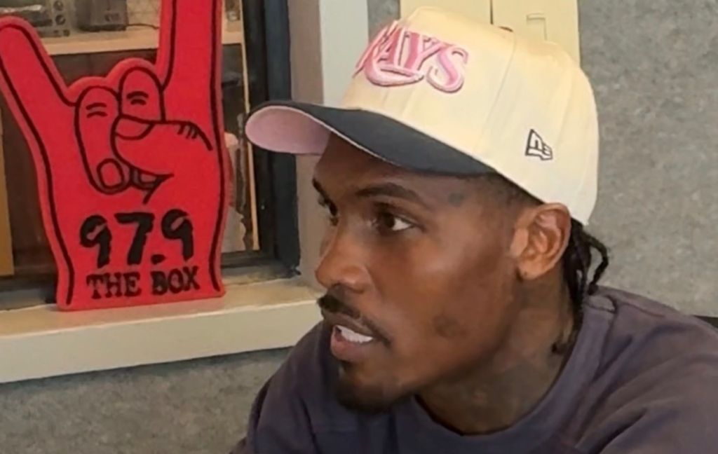 A man wearing a New Era baseball cap and a dark jacket, standing in front of a sign that says "97.9 THE BOX".