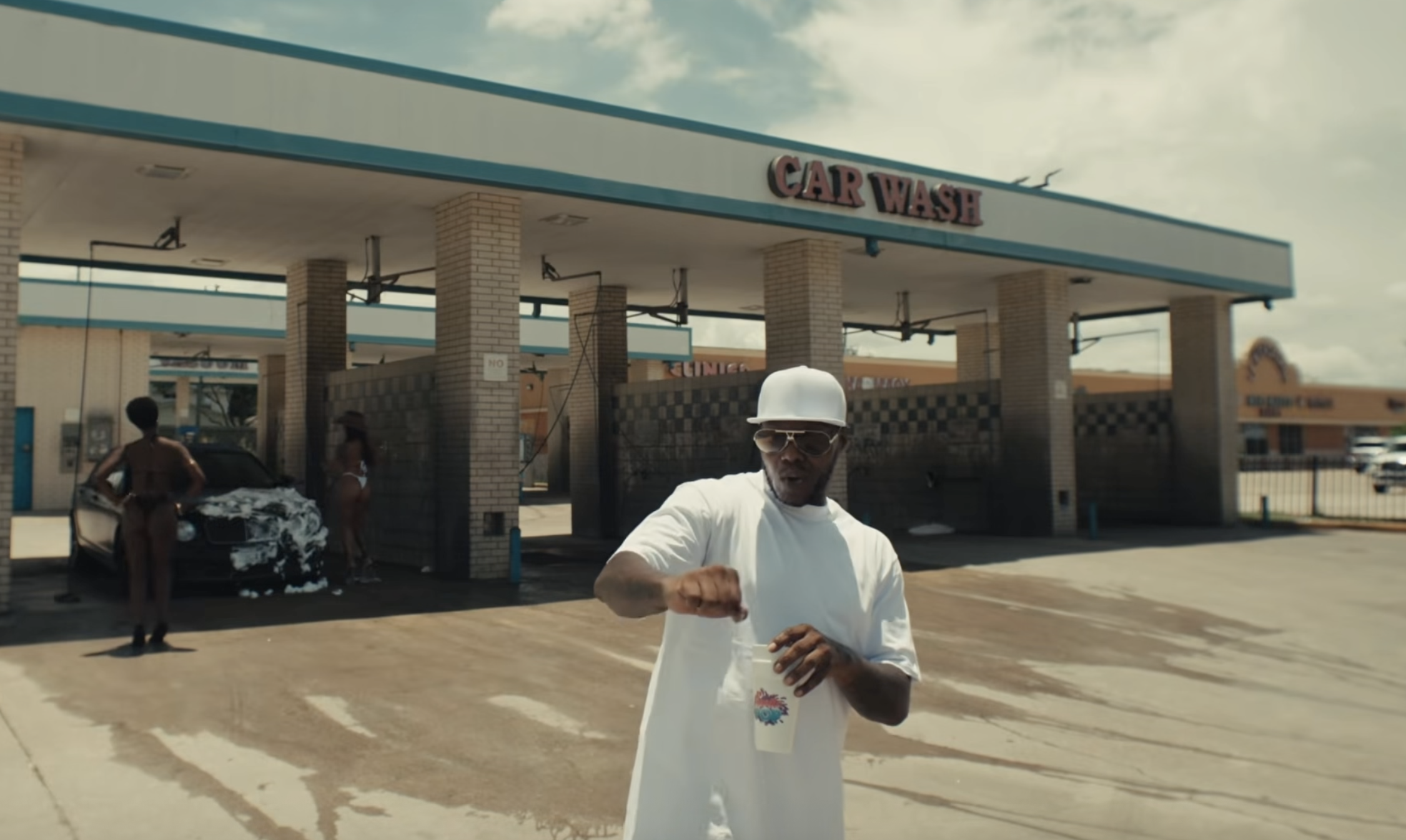 A person in a white outfit and hard hat stands in front of a car wash facility with the "Car Wash" sign visible.