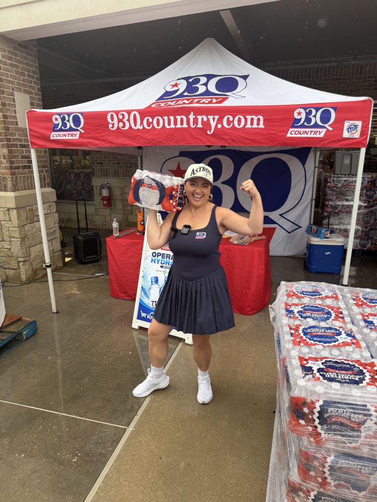 A smiling woman wearing an Astros cap and tank top stands in front of a 93Q Country tent, surrounded by promotional items.