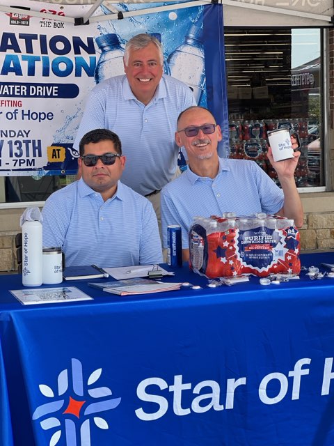 Three men standing behind a Star of Hope booth, one holding a mobile device and another holding a box of supplies, with a large banner advertising a "Water Drive" event.