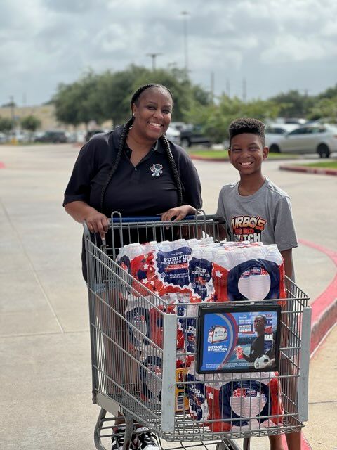 A smiling woman and young boy standing in front of a shopping cart filled with various items, including a Walmart logo visible on the cart.