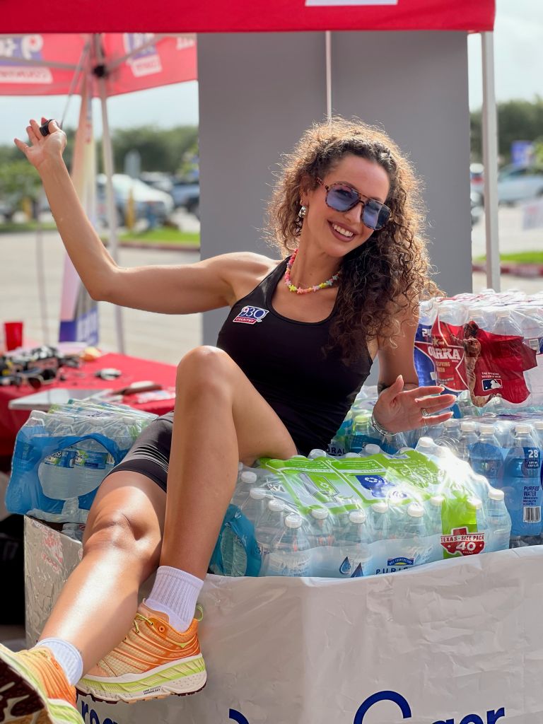 A smiling woman wearing sunglasses and a black tank top sits among cases of bottled water and other supplies, gesturing with her arms.