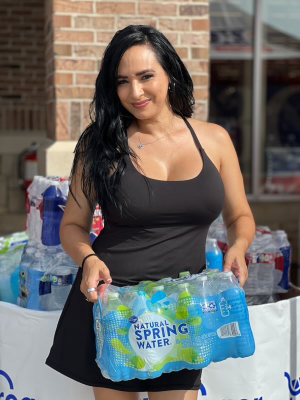 A woman in a black tank top holding a case of Natural Spring Water bottles.
