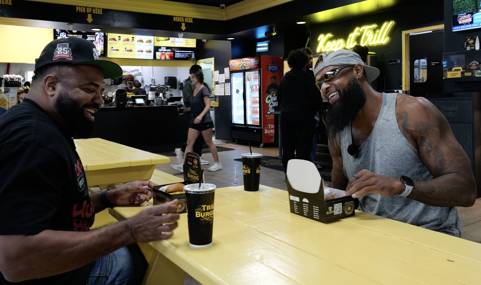 Two men smiling and enjoying food and drinks at a fast food restaurant called "Trillburger" with neon "Keep it Chill" sign visible.