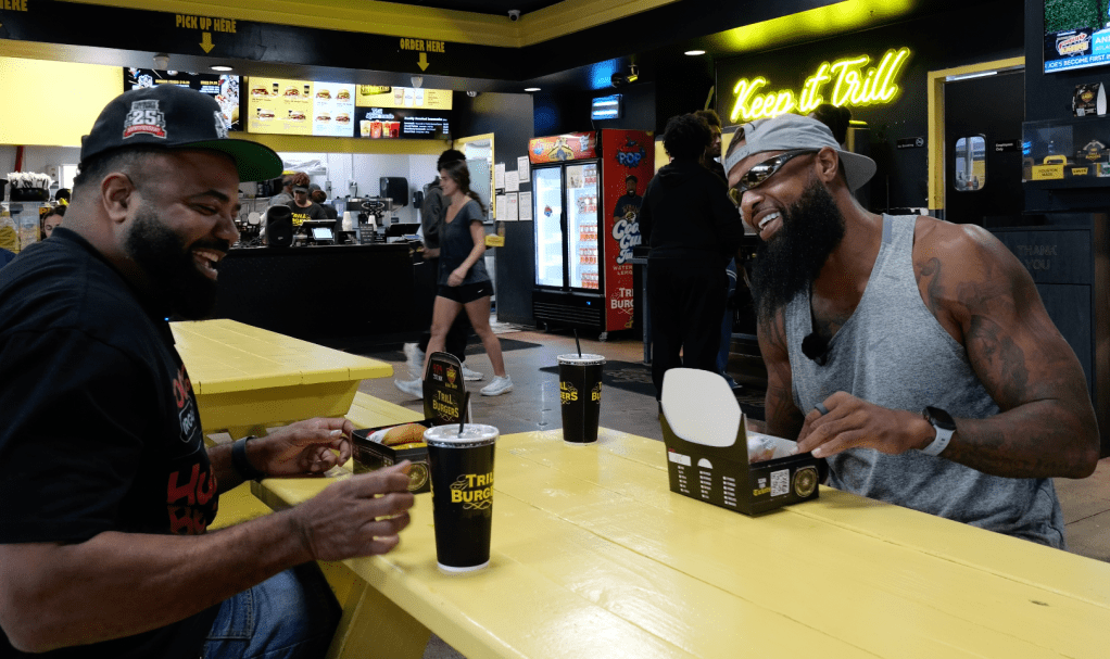 Two men smiling and enjoying food and drinks at a fast food restaurant called "Trillburger" with neon "Keep it Chill" sign visible.