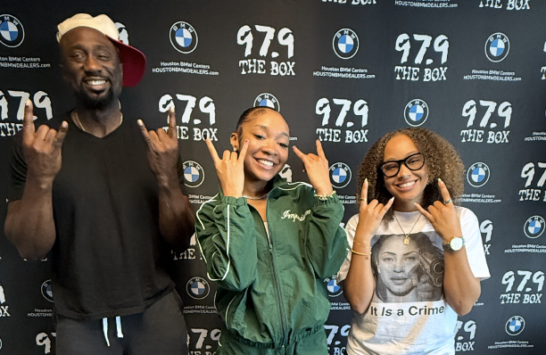 Three people making peace signs in front of a backdrop with "979 THE BOX" and BMW logos.