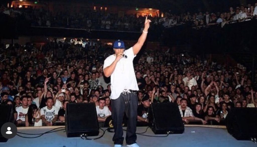 A performer on stage addressing a large, enthusiastic crowd at a live event. The performer is wearing a white shirt and baseball cap.