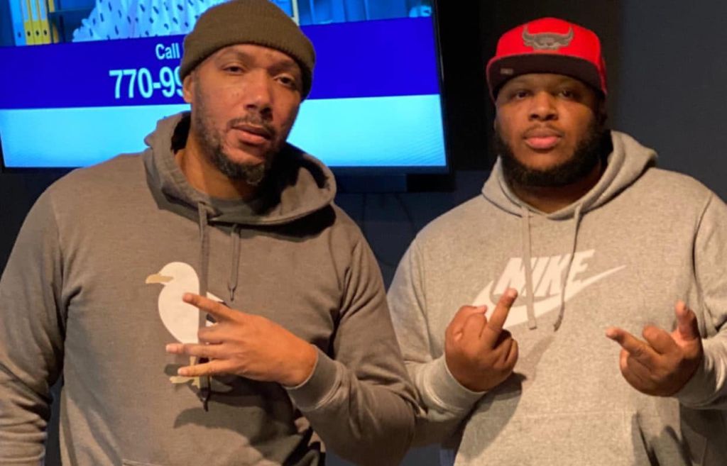 Two men wearing hoodies and hats, one with a red hat and the other with a brown hat, standing in front of a TV screen showing a phone number.