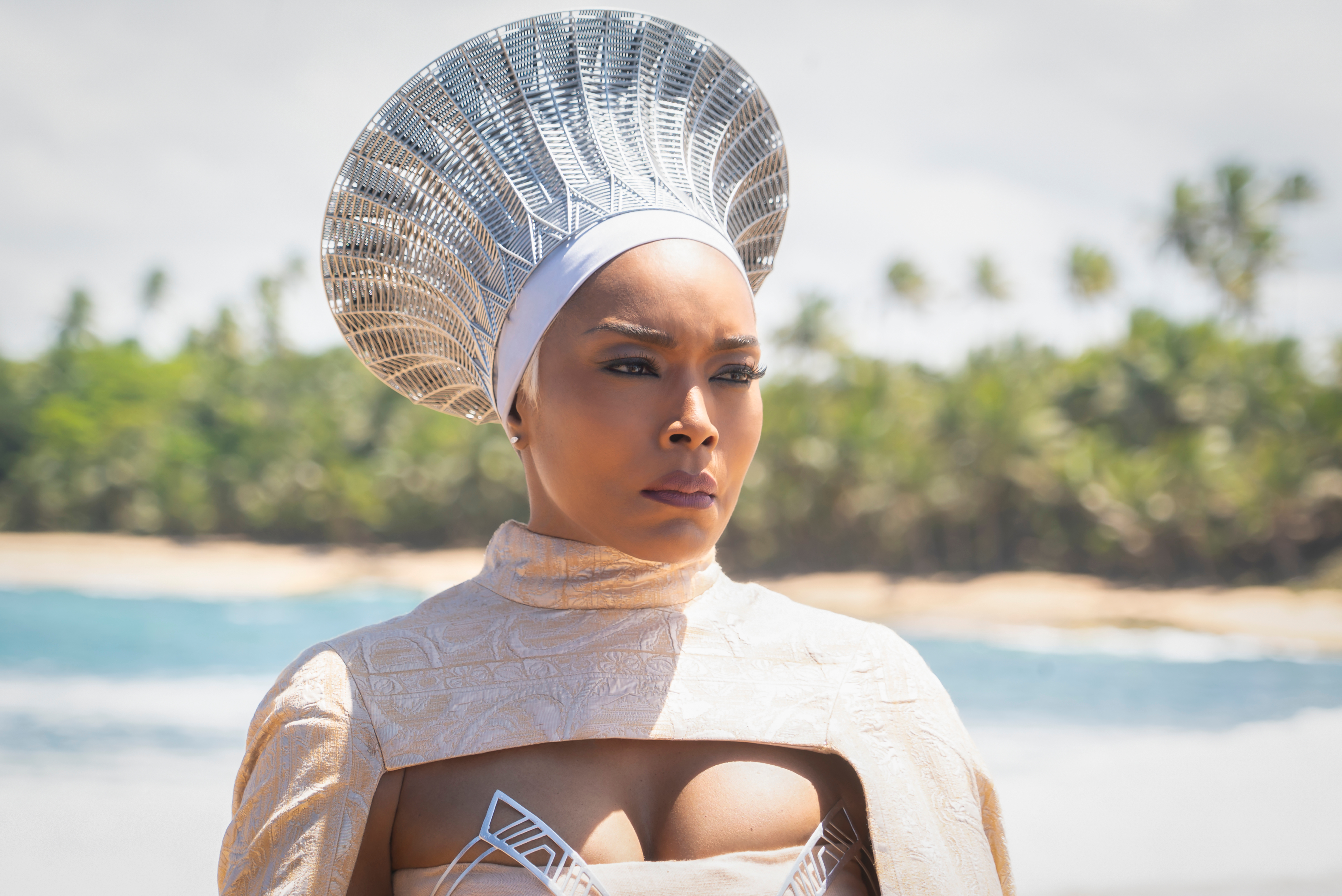 A woman wearing an elaborate headdress and a white costume stands on a beach, looking serious.