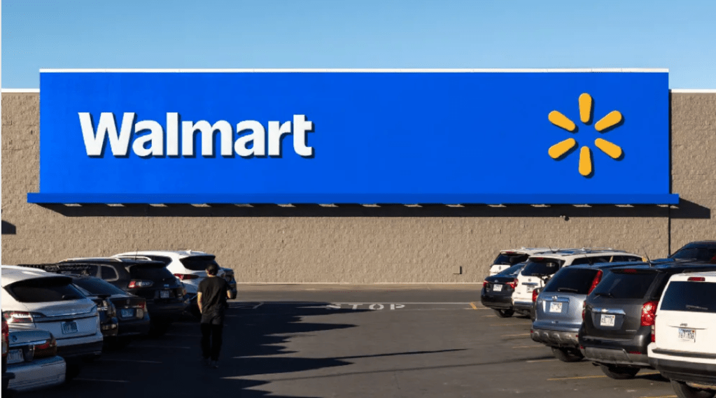 Walmart store exterior with large blue and yellow logo sign, parking lot with several vehicles.