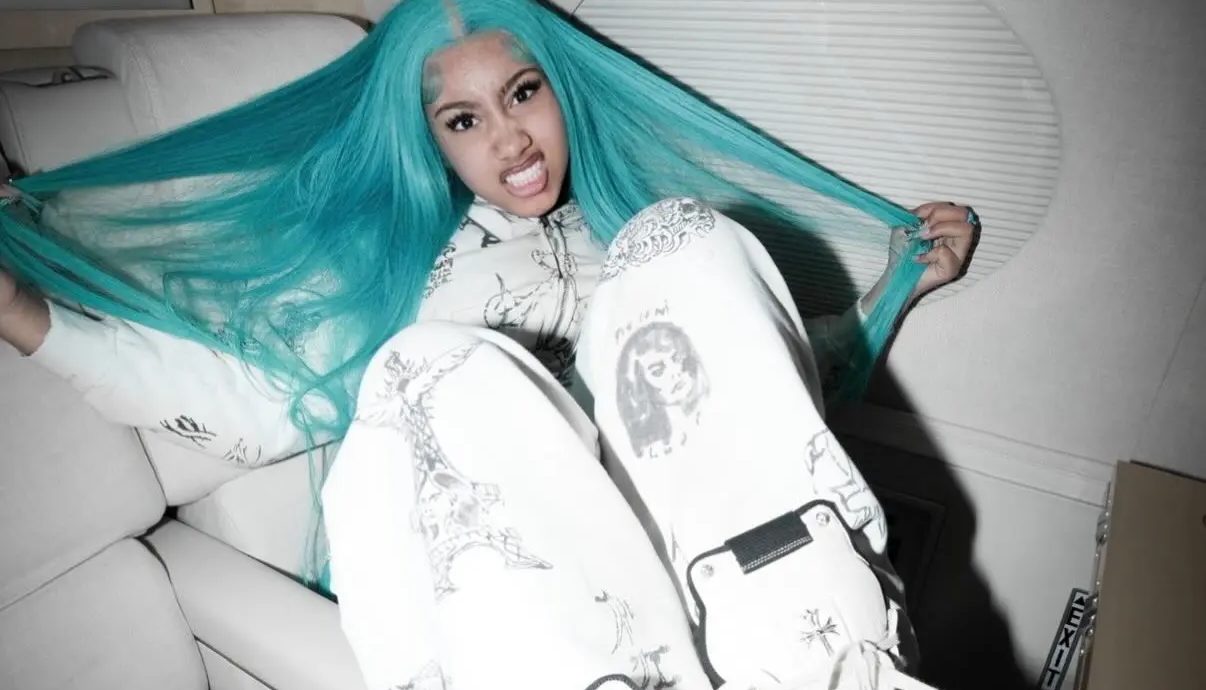 A person with long teal-colored hair wearing a white jacket with printed designs, making an expressive facial expression.