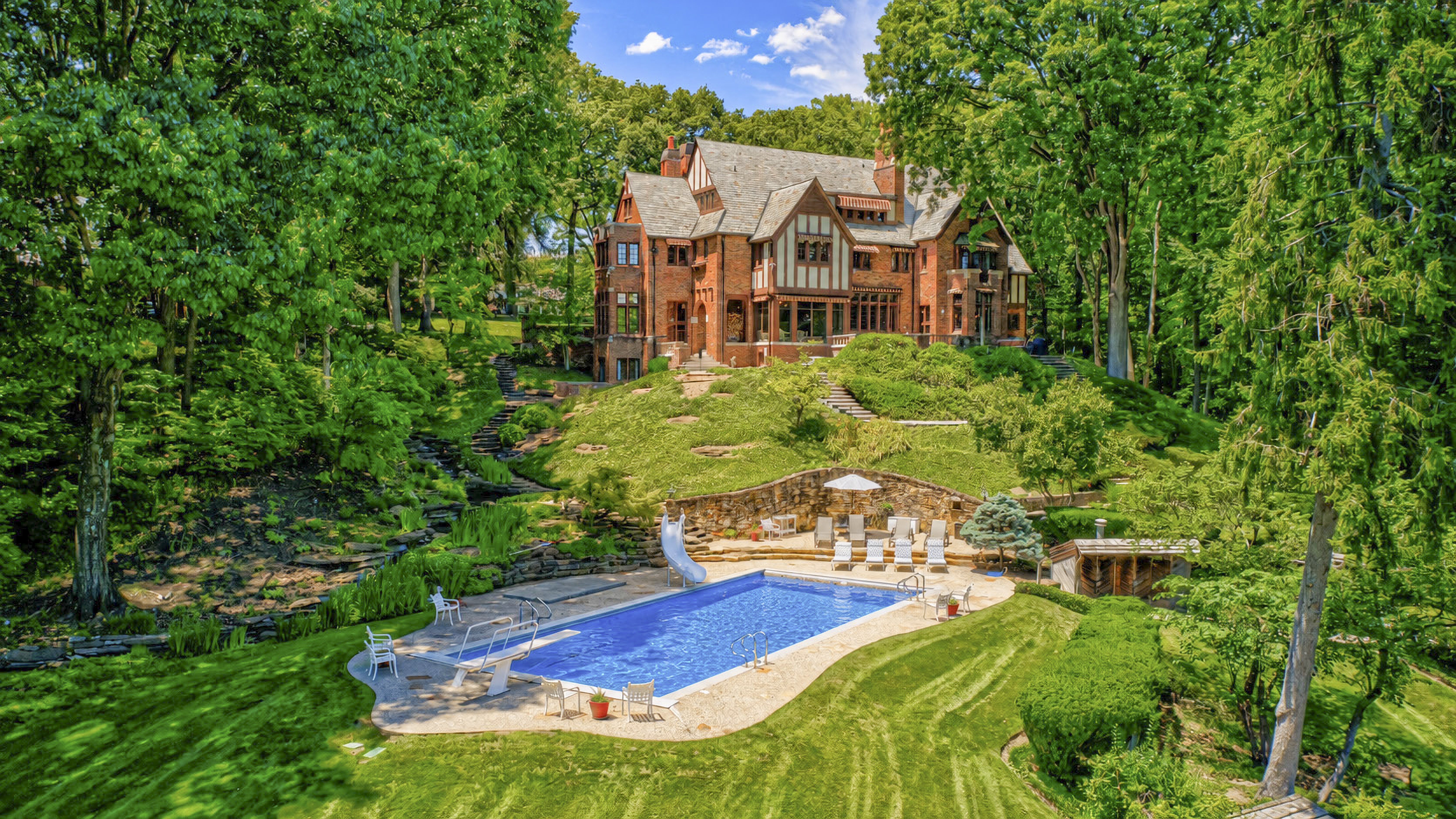 A large, ornate Tudor-style mansion surrounded by lush, verdant gardens and a swimming pool in a tranquil, wooded setting.