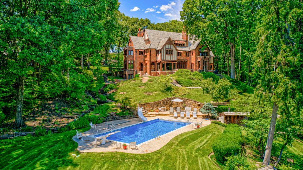 A large, ornate Tudor-style mansion surrounded by lush, verdant gardens and a swimming pool in a tranquil, wooded setting.