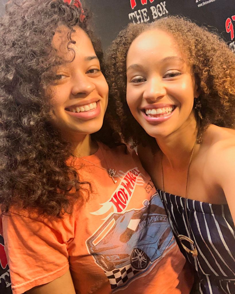 Two smiling young women with curly hair, one wearing an orange t-shirt with a car graphic and the other wearing a striped top.