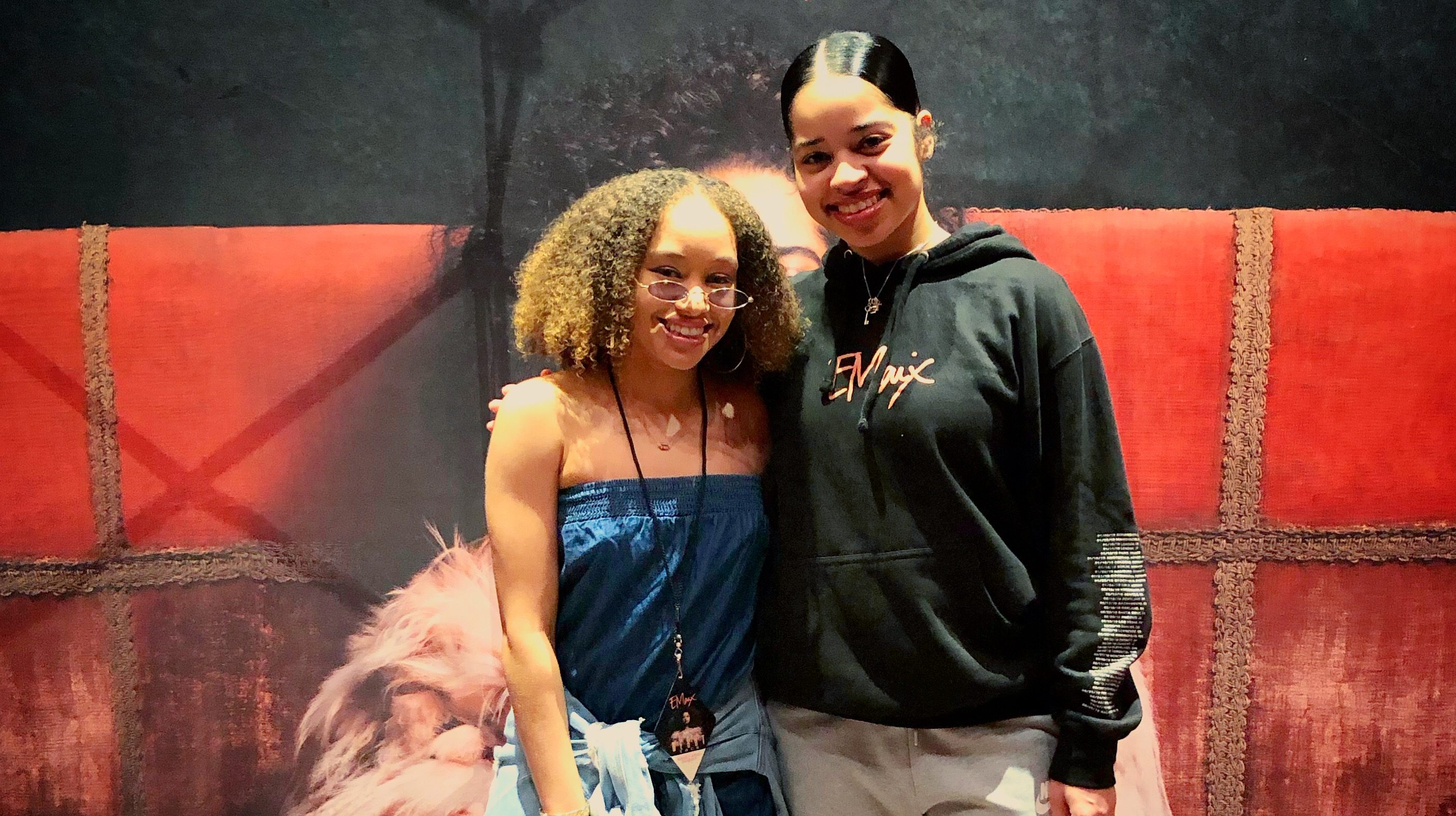 Two young women smiling and posing together in front of a red and black backdrop with a large feathered decoration.