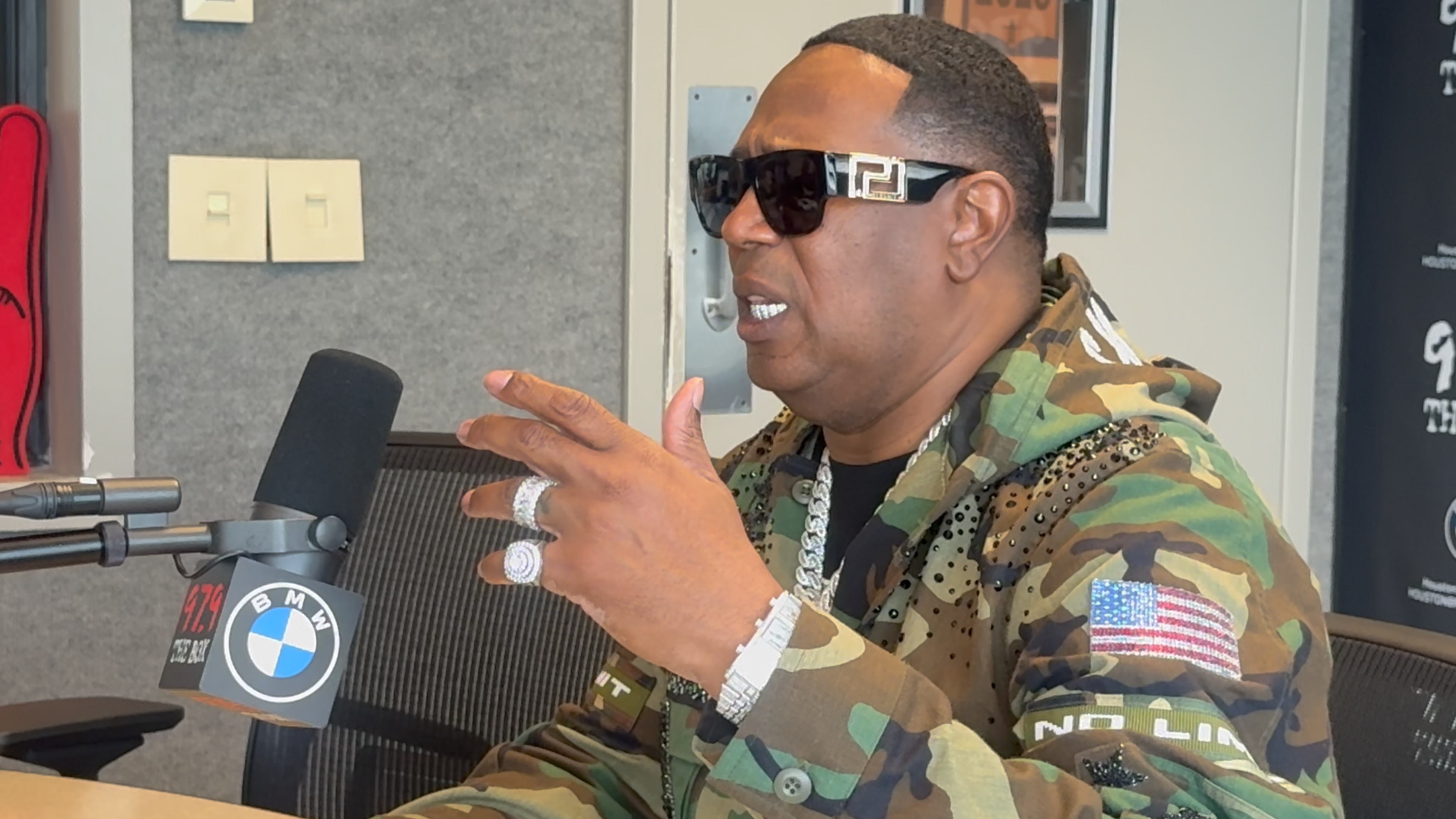 A man in camouflage military uniform wearing sunglasses and jewelry, speaking into a microphone in a studio setting.