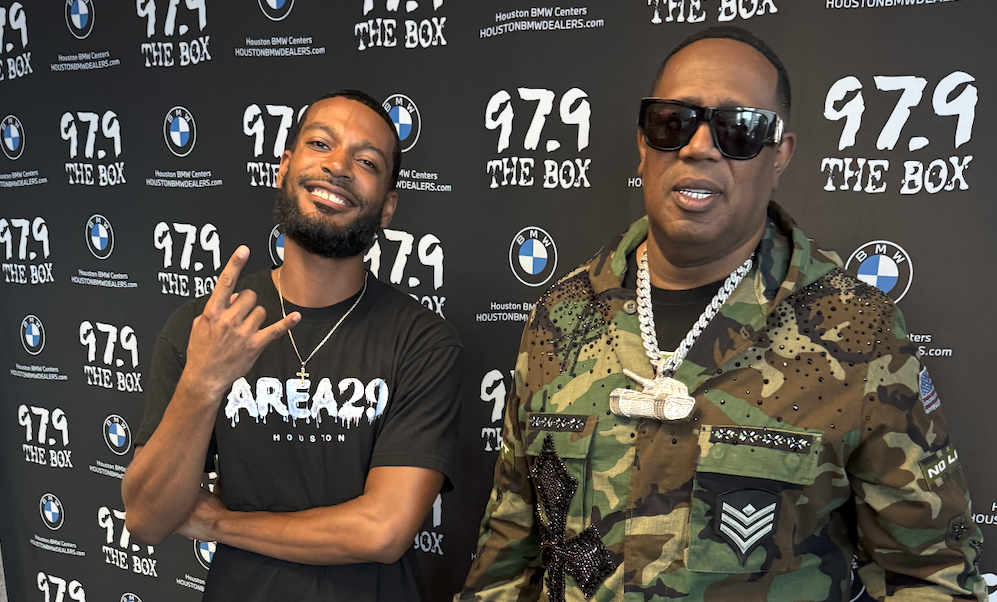 Two men posing in front of a backdrop with "97.9 THE BOX" and BMW logos, one wearing a camouflage outfit and the other wearing a black t-shirt with "AREA 20" printed on it.