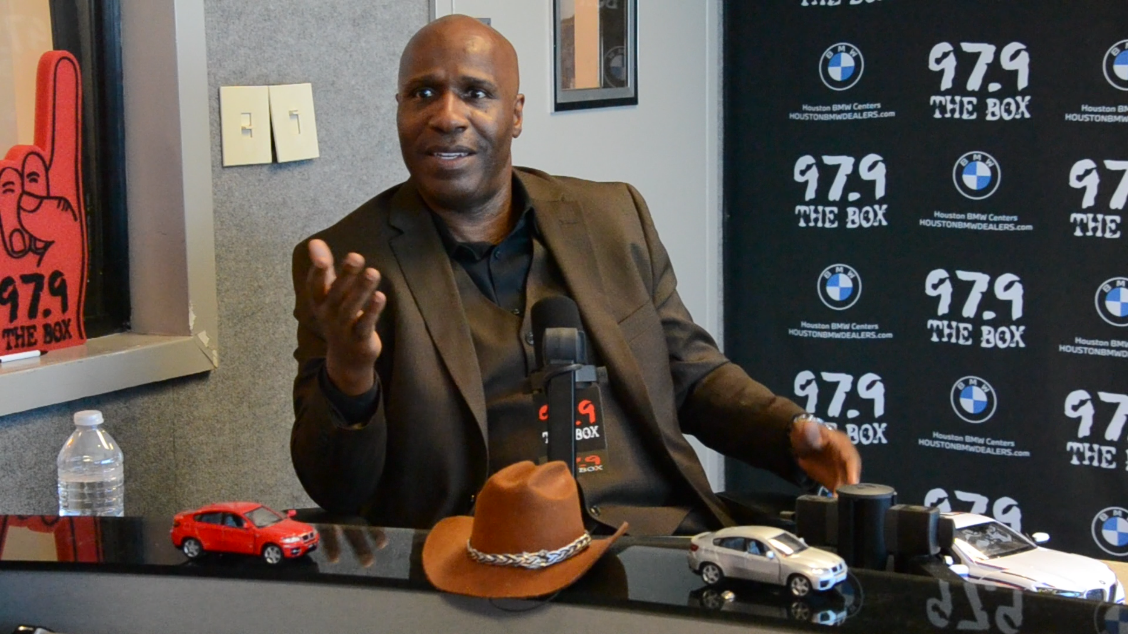 A man wearing a suit and hat sitting at a desk with toy cars and BMW-branded radio station signage behind him.