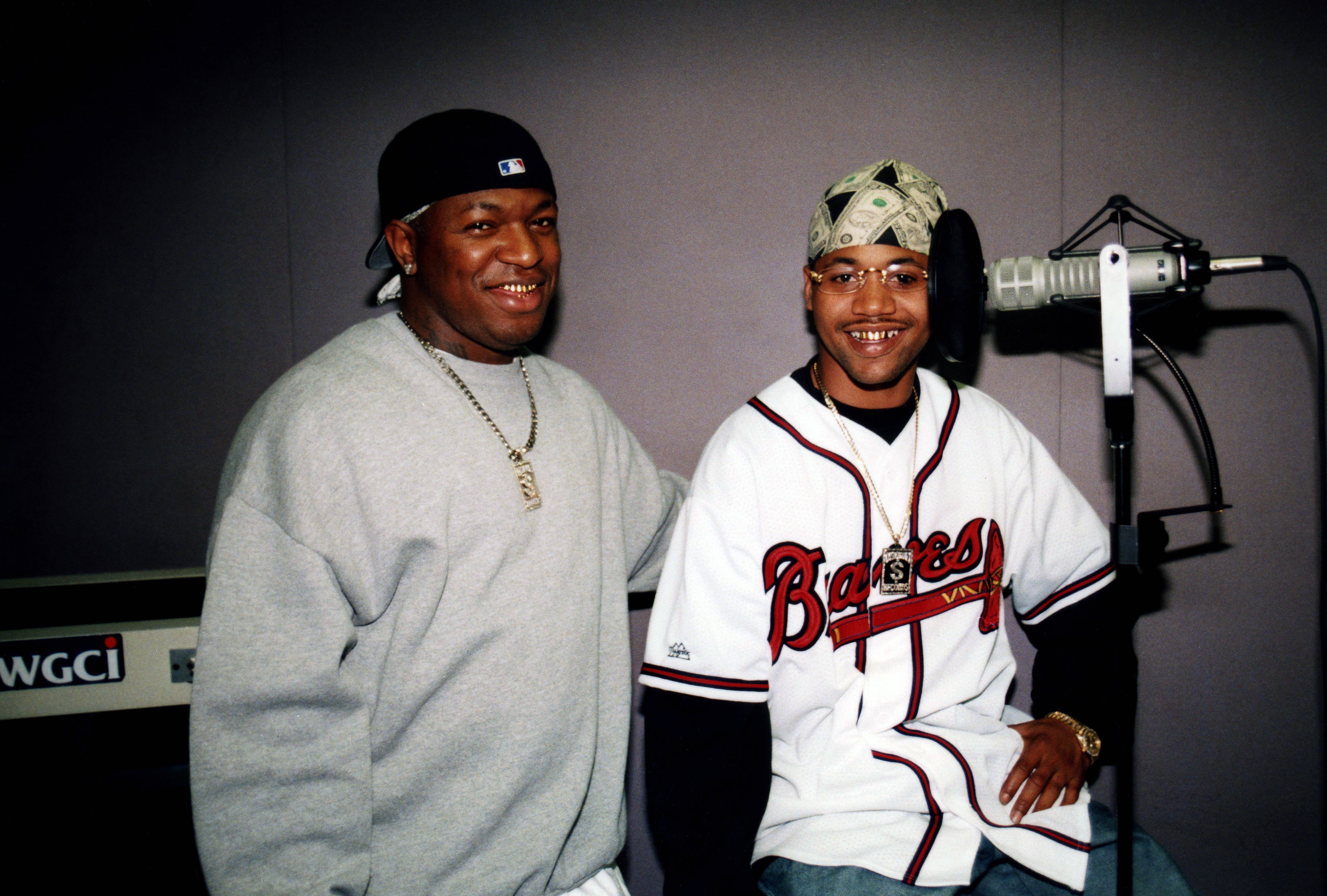 Two smiling African American men, one wearing a baseball cap and the other a bandana, posing together in a radio studio.